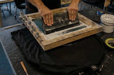 Hands pulling a squeegee across a silkscreen frame over a blank black t-shirt